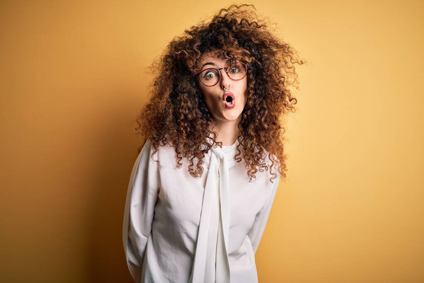 Young beautiful brunette woman with curly hair and piercing wearing shirt and glasses afraid and shocked with surprise expression, fear and excited face. - Photo, Image