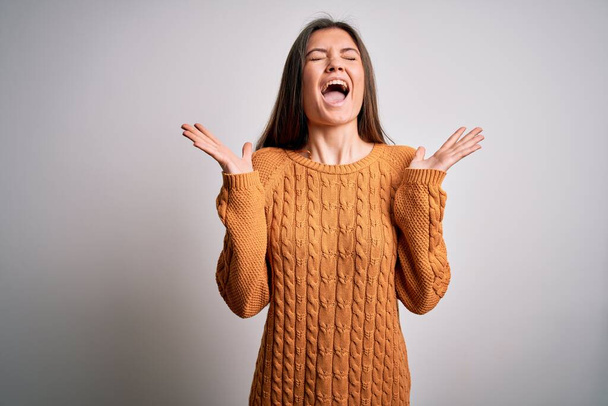 Young beautiful woman with blue eyes wearing casual sweater standing over white background celebrating mad and crazy for success with arms raised and closed eyes screaming excited. Winner concept - Fotoğraf, Görsel