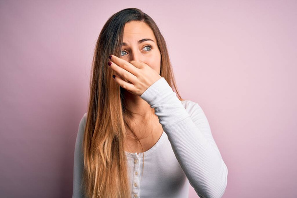 Young beautiful blonde woman with blue eyes wearing white t-shirt over pink background smelling something stinky and disgusting, intolerable smell, holding breath with fingers on nose. Bad smell - Valokuva, kuva