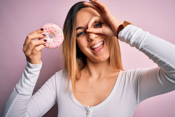 Jovem mulher loira bonita com olhos azuis segurando rosa donut sobre fundo isolado com rosto feliz sorrindo fazendo ok sinal com a mão no olho olhando através dos dedos
 - Foto, Imagem