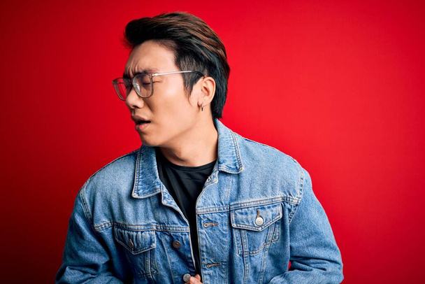 Young handsome chinese man wearing denim jacket and glasses over red background with hand on stomach because nausea, painful disease feeling unwell. Ache concept. - Foto, immagini