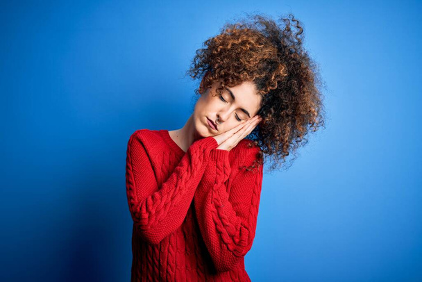 Молодая красивая женщина с вьющимися волосами и пирсинг носить случайные красный свитер спит усталый сон и позировать с руками вместе, улыбаясь с закрытыми глазами
. - Фото, изображение
