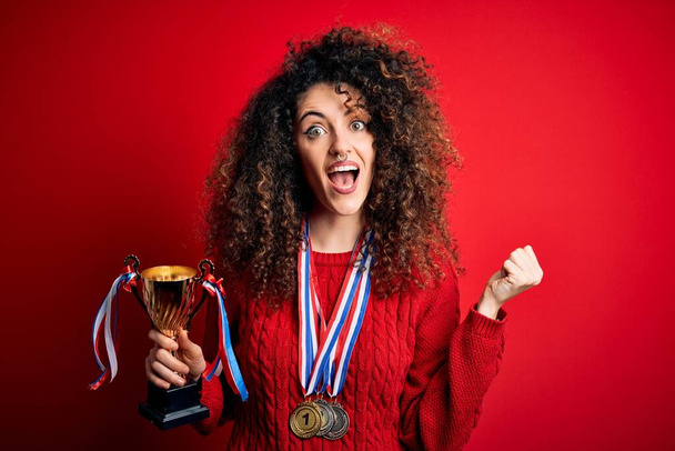 Young beautiful successful woman with curly hair and piercing holding trophy wearing medals screaming proud and celebrating victory and success very excited, cheering emotion - Photo, Image