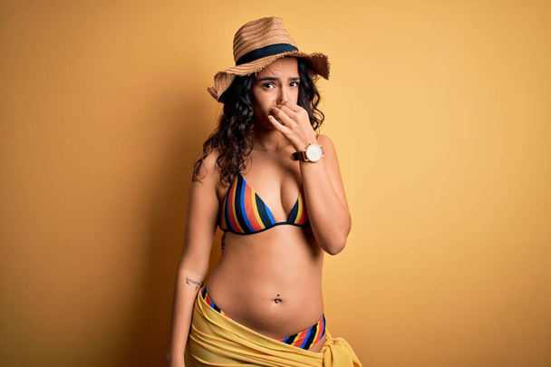 Young beautiful woman with curly hair on vacation wearing bikini and summer hat smelling something stinky and disgusting, intolerable smell, holding breath with fingers on nose. Bad smell - Photo, image