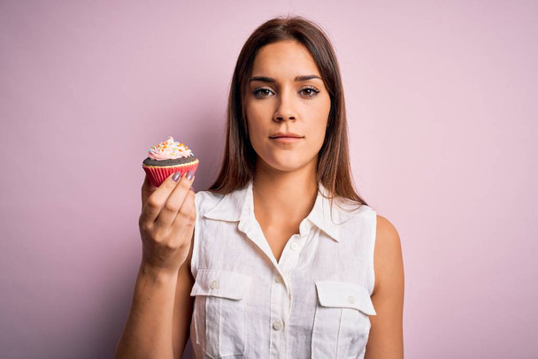 若いです美しいですブルネット女性食べるチョコレートカップケーキ上の隔離されたピンクの背景とともに自信を持って式上のスマート顔考え深刻な - 写真・画像