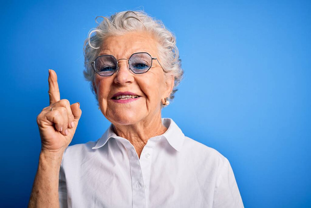 Senior belle femme portant chemise élégante et des lunettes sur fond bleu isolé pointant doigt vers le haut avec idée réussie. Sorti et heureux. Numéro un
. - Photo, image