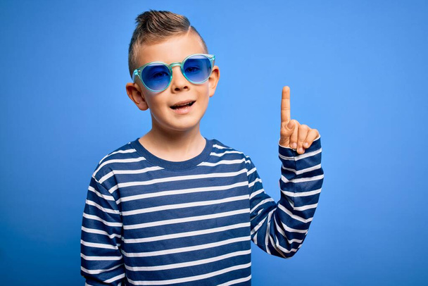 Νεαρό μικρό καυκάσιο παιδί με μπλε μάτια στέκεται φορώντας γυαλιά ηλίου πάνω από το μπλε φόντο δείχνοντας το δάχτυλο επάνω με την επιτυχή ιδέα. Έξοδος και ευτυχία. Νούμερο ένα.. - Φωτογραφία, εικόνα