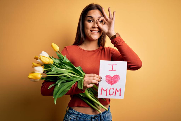 Beautiful woman celebrating mothers day holding love mom message and bouquet of tulips with happy face smiling doing ok sign with hand on eye looking through fingers - Foto, Bild