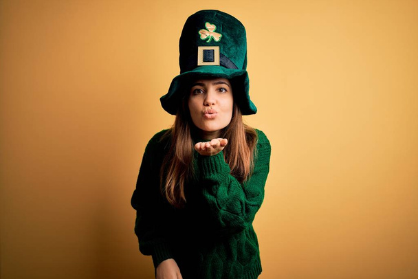 Молодая красивая брюнетка в зеленой шляпе на праздновании дня святого Патрика смотрит в камеру и целует рукой в воздух, будучи красивой и сексуальной. Любовное выражение
. - Фото, изображение