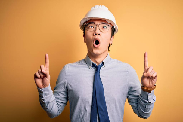 Young handsome chinese architect man wearing safety helmet and tie over yellow background amazed and surprised looking up and pointing with fingers and raised arms. - Foto, imagen