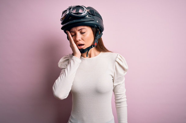 Jovem bela mulher motociclista com olhos azuis usando capacete de moto sobre fundo rosa bocejo cansado cobrindo metade do rosto, olho e boca com a mão. Cara dói na dor
. - Foto, Imagem