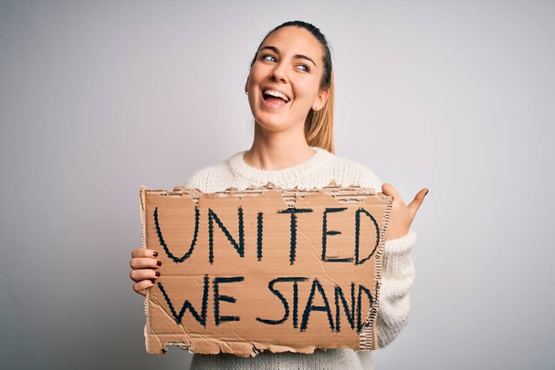 Young beautiful blonde woman with blue eyes asking for united stand holding banner pointing and showing with thumb up to the side with happy face smiling - Fotografie, Obrázek