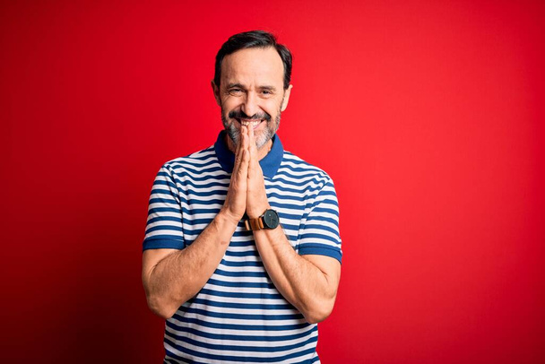 Middle age hoary man wearing casual striped polo standing over isolated red background praying with hands together asking for forgiveness smiling confident. - Photo, Image