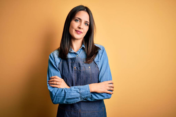 Young baker woman with blue eyes wearing apron standing over isolated yellow background happy face smiling with crossed arms looking at the camera. Positive person. - Φωτογραφία, εικόνα