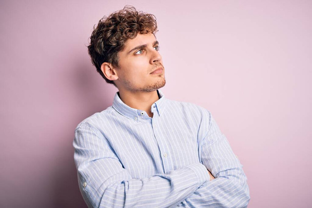 Young blond handsome man with curly hair wearing striped shirt over white background looking to the side with arms crossed convinced and confident - Фото, зображення