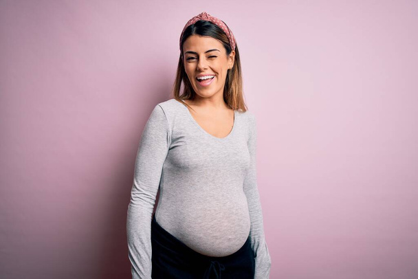 Νεαρή όμορφη μελαχρινή γυναίκα έγκυος περιμένει μωρό πάνω από απομονωμένο ροζ φόντο κλείνει το μάτι κοιτάζοντας την κάμερα με σέξι έκφραση, χαρούμενο και χαρούμενο πρόσωπο. - Φωτογραφία, εικόνα