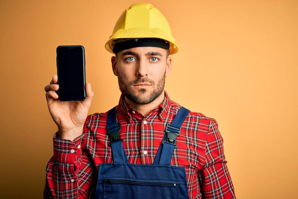Молодой строитель в защитном шлеме показывает экран смартфона на жёлтом фоне с уверенным выражением на умном лице, думающем серьезно
 - Фото, изображение