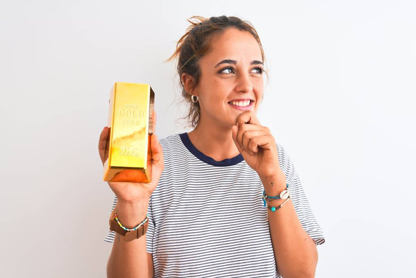 Jovem mulher ruiva bonita segurando lingote de ouro sobre fundo isolado rosto sério pensando em questão, ideia muito confusa
 - Foto, Imagem