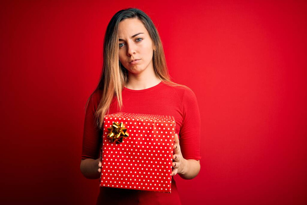 Young beautiful blonde woman with blue eyes holding birthday gift over red background with a confident expression on smart face thinking serious - Fotó, kép