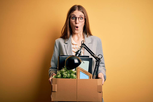 Young beautiful redhead businesswoman fired holding carboard box over yellow background scared in shock with a surprise face, afraid and excited with fear expression - 写真・画像