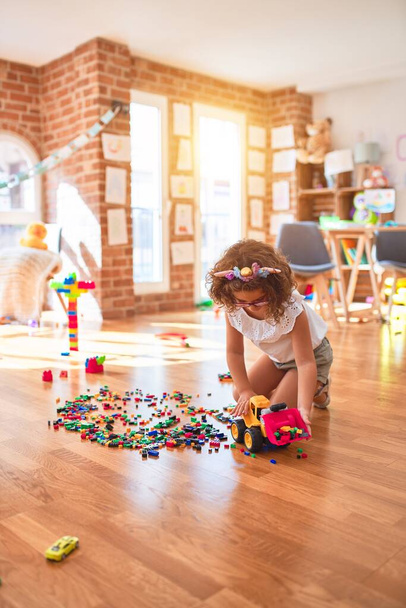 Beautiful toddler wearing glasses and unicorn diadem playing with tractor and building blocks at kindergarten - Photo, Image
