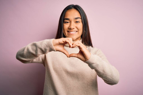 Joven hermosa mujer asiática con suéter casual cuello alto sobre fondo rosa sonriendo en el amor haciendo forma de símbolo del corazón con las manos. Concepto romántico
. - Foto, imagen