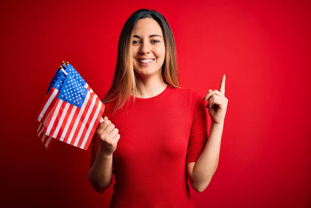 Junge schöne blonde patriotische Frau mit blauen Augen, die die Flaggen der Vereinigten Staaten hält, überrascht mit einer Idee oder Frage, die mit einem glücklichen Gesicht zeigt, Nummer eins - Foto, Bild