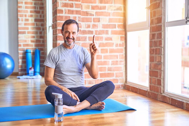 Középkorú jóképű sportember ül szőnyegen csinál nyújtás jóga gyakorlat tornaterem mutatva ujját fel a sikeres ötlet. Kilépett és boldog. Egyes számú.. - Fotó, kép
