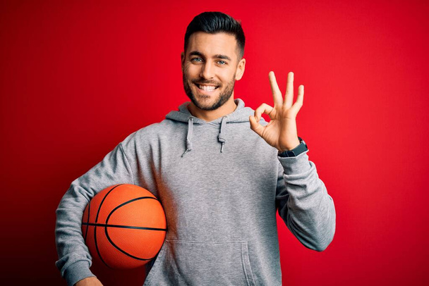 Young sports man holding basketball ball over red isolated background doing ok sign with fingers, excellent symbol - Foto, immagini