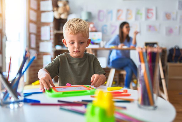 Genç beyaz çocuk öğretmeniyle oyun okulunda oynuyor. Anne ve oğlu oyun odasında renkli kalemlerle çizim yapıyor. Arka planda oturan genç kadın da masada oturuyor.. - Fotoğraf, Görsel
