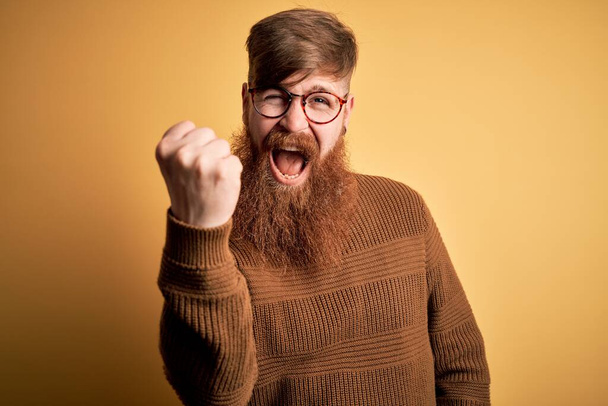 Красивый ирландский рыжий мужчина с бородой в очках и зимнем свитере на жёлтом фоне злой и безумный, поднимающий кулак, раздражённый и разъяренный, крича от гнева. Ярость и агрессивность
. - Фото, изображение