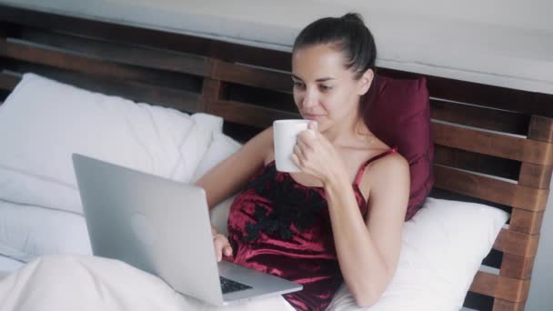 woman works on laptop drinking coffee in comfortable bed - Záběry, video