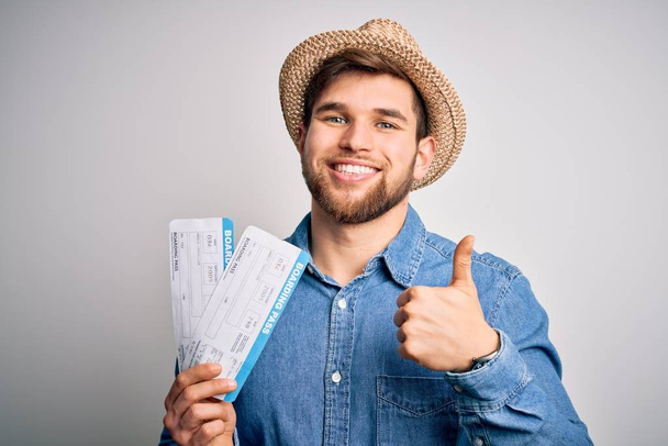 Young blond tourist man with blue eyes on vacation wearing hat holding boarding pass happy with big smile doing ok sign, thumb up with fingers, excellent sign - Foto, immagini