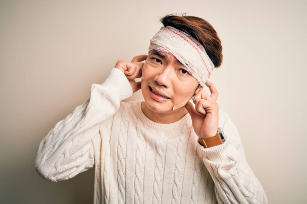 Jonge knappe Chinese man gewond door ongeval dragen van verband en strips op het hoofd bedekt oren met vingers met geïrriteerde uitdrukking voor het lawaai van luide muziek. Dovemansconcept. - Foto, afbeelding