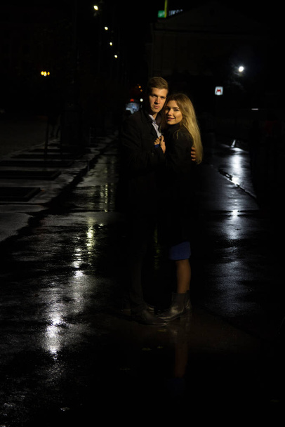 ζευγάρι στην αγάπη αγκαλιάζει σε ένα σκοτεινό φόντο, νύχτα, βροχή, κτίρια στην πόλη, ένα πάρκο, χαμογελώντας, έλξη. Ο τύπος αγκαλιάζει το κορίτσι. Ημέρα του Αγίου Βαλεντίνου σκοτεινό δρόμο και βροχή - Φωτογραφία, εικόνα