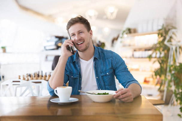 Mladý pohledný usměvavý muž pije horkou kávu a jí salát k obědu, zatímco mluví na svém smartphonu během přestávky v kavárně, multitasking koncept. - Fotografie, Obrázek