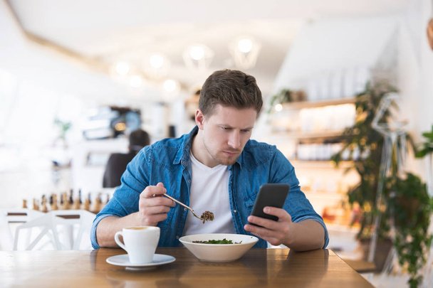 Young handsome smiling man drinks his hot coffee and eats salad for lunch while typing message in his smartphone during break at cafe , multitasking concept. - Zdjęcie, obraz