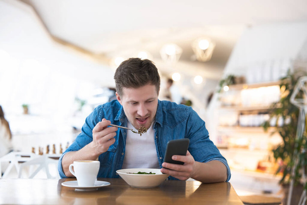 Νεαρός όμορφος χαμογελαστός άντρας πίνει τον ζεστό του καφέ και τρώει σαλάτα για μεσημεριανό ενώ διαβάζει το μήνυμα στο smartphone του κατά τη διάρκεια του διαλείμματος στο cafe, κάνοντας πολλές δουλειές. - Φωτογραφία, εικόνα
