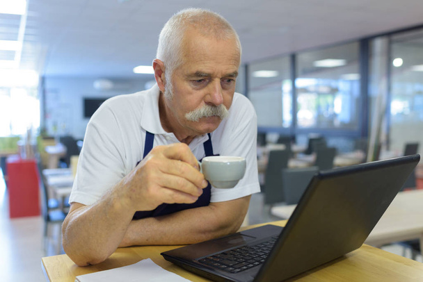elderly cafeteria worker drinking coffee readin laptop - Zdjęcie, obraz