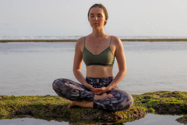 Junge Frau sitzt auf dem Felsen in Lotus-Pose, meditiert, praktiziert Yoga und Pranayama. Tiefer Atem und Konzentration mit geschlossenen Augen. Outdoor-Yoga am Strand. Melasti Strand, Bali, Indonesien - Foto, Bild
