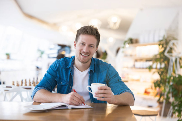young handsome smiling man drinks coffee during his lunch break in the cafe taking notes to his planner looking busy, multitasking concept. - Photo, image