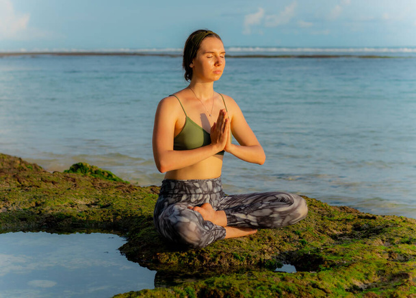 Junge Frau, meditiert, praktiziert Yoga und Pranayama am Strand. Yoga bei Sonnenuntergang. Hände im namaste mudra, geschlossene Augen und Konzentration. Lotus Pose. Melasti Strand, Bali, Indonesien - Foto, Bild