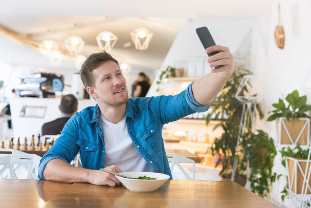 joven guapo sonriente hombre disparando selfie en su teléfono inteligente mientras come ensalada durante su hora de almuerzo en el café, concepto multitarea
. - Foto, imagen