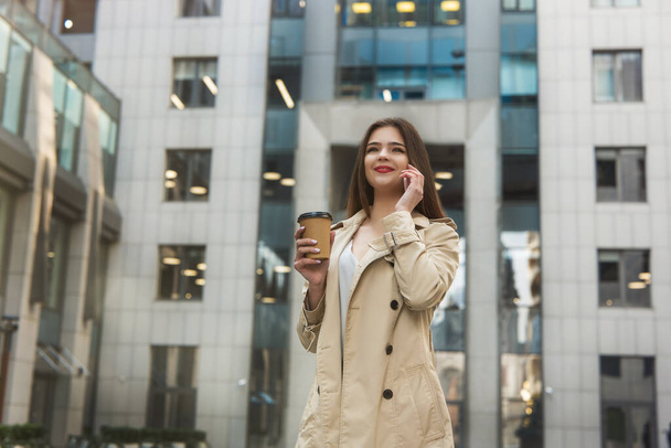 Joven mujer de negocios bebiendo café caliente en el camino en medio de la calle cerca del centro de oficinas hablando en su teléfono mirando feliz, concepto multitarea
. - Foto, imagen
