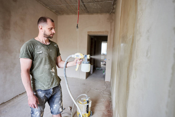 Man bouwer is schilderen met airbrush muur, huis reparatie. Op de achtergrond is veel werk te zien. Een man met een baard is gekleed in met verf besmeurde kleding. Reparatieconcept - Foto, afbeelding