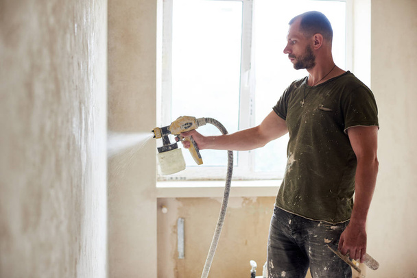 Man builder is painting with airbrush wall, house repairment, against window in the daytime. Man with a beard is dressed in paint-smeared T-shirt. Repair concept - Photo, Image