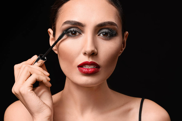 Beautiful young woman applying makeup against dark background - Photo, Image