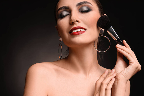 Beautiful young woman applying makeup against dark background - Photo, image