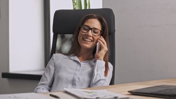 Happy businesswoman wearing eyeglasses talking by smartphone and relaxing on office chair in office - Felvétel, videó