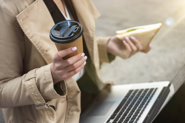 mujer de negocios sosteniendo el ordenador portátil de rodillas con una taza de café y un sándwich en las manos sentada cerca de la oficina durante el almuerzo, comiendo sobre la marcha
 . - Foto, imagen